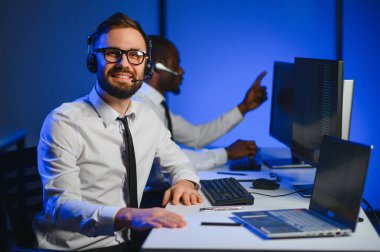 Mutlu Güvenilir Teknik Müşteri Destek Uzmanı Meslektaşlar ve Ekranlarla Dolu Gözetleme ve Kontrol Odasında Kulaklık Çağrısı Yapıyor.