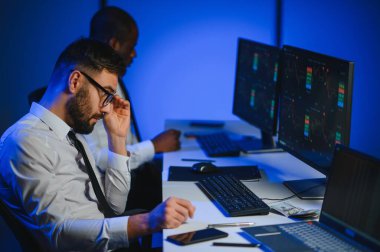 İki işadamı, finans analisti ya da yatırım danışmanı ellerinde polis bilgisayarları ve belgelerle büro masasında oturuyorlar.