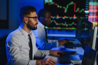 İki işadamı, finans analisti ya da yatırım danışmanı ellerinde polis bilgisayarları ve belgelerle büro masasında oturuyorlar.