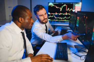 İki işadamı, finans analisti ya da yatırım danışmanı ellerinde polis bilgisayarları ve belgelerle büro masasında oturuyorlar.