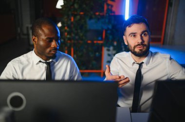 İki işadamı, finans analisti ya da yatırım danışmanı ellerinde polis bilgisayarları ve belgelerle büro masasında oturuyorlar.