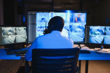 Telsizle konuşurken masaüstü bilgisayar ekranına bakan Afrikalı Amerikalı güvenlik görevlisi.