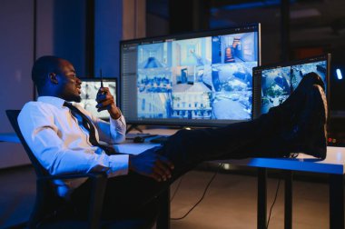 Ciddi bir Afrikalı Amerikalı güvenlik görevlisi güvenlik odasında güvenlik kamerası görüntülerini izlerken bilgisayar ekranından otel hayatını izliyor.