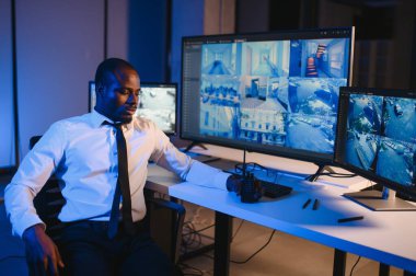 Ciddi bir Afrikalı Amerikalı güvenlik görevlisi güvenlik odasında güvenlik kamerası görüntülerini izlerken bilgisayar ekranından otel hayatını izliyor.