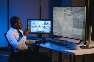 Telsizle konuşurken masaüstü bilgisayar ekranına bakan Afrikalı Amerikalı güvenlik görevlisi.