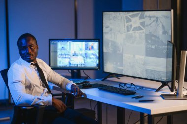 Ciddi bir Afrikalı Amerikalı güvenlik görevlisi güvenlik odasında güvenlik kamerası görüntülerini izlerken bilgisayar ekranından otel hayatını izliyor.