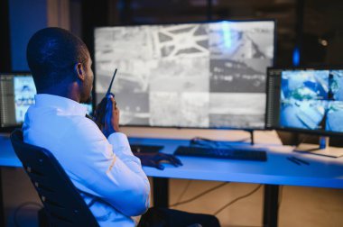 Ciddi bir Afrikalı Amerikalı güvenlik görevlisi güvenlik odasında güvenlik kamerası görüntülerini izlerken bilgisayar ekranından otel hayatını izliyor.