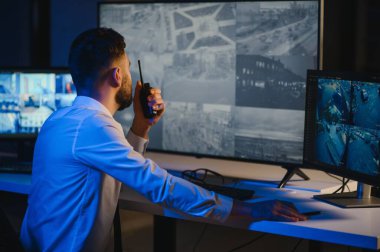 Erkek güvenlik görevlisi, bilgisayardaki birden fazla CCTV görüntüsünü izlerken telsizden konuşuyor..