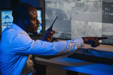 Ciddi bir Afrikalı Amerikalı güvenlik görevlisi güvenlik odasında güvenlik kamerası görüntülerini izlerken bilgisayar ekranından otel hayatını izliyor.