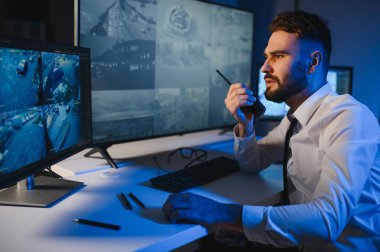 Erkek güvenlik görevlisi, bilgisayardaki birden fazla CCTV görüntüsünü izlerken telsizden konuşuyor..