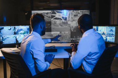 İki güvenlik görevlisi iş yerinde bilgisayar monitörlerinin önünde oturuyorlar ve monitörlerdeki güvenlik kameralarını izliyorlar.