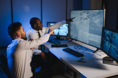 İki güvenlik görevlisi iş yerinde bilgisayar monitörlerinin önünde oturuyorlar ve monitörlerdeki güvenlik kameralarını izliyorlar.