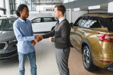 Başarılı bir anlaşmadan sonra gülümseyen araba satıcısı müşteriyle tokalaşıyor, bayilik merkezinde yeni bir araç alan mutlu Afrikalı Amerikalı adam, modern oto galerisindeki satış elemanıyla anlaşma yapıyor.