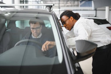 Erkek araba satıcısı, galeride dalgın genç bir müşteriyle sohbet ediyor. Satış merkezinden araba alırken adam dinliyor.