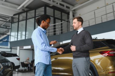 Başarılı bir anlaşmadan sonra gülümseyen araba satıcısı müşteriyle tokalaşıyor, bayilik merkezinde yeni bir araç alan mutlu Afrikalı Amerikalı adam, modern oto galerisindeki satış elemanıyla anlaşma yapıyor.