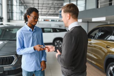 Başarılı bir anlaşmadan sonra gülümseyen araba satıcısı müşteriyle tokalaşıyor, bayilik merkezinde yeni bir araç alan mutlu Afrikalı Amerikalı adam, modern oto galerisindeki satış elemanıyla anlaşma yapıyor.