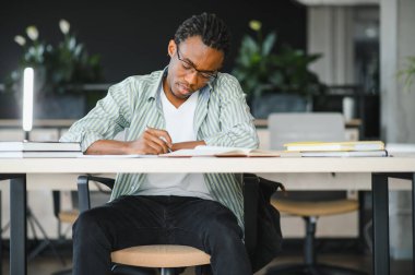 Genç adam ders kitabından notlar alıyor, üniversite kütüphanesinde sınav hazırlıyor, odaklanmış öğrenim ve öğrenim görüyor.