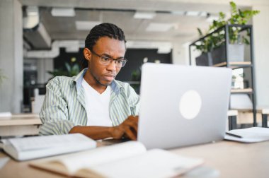 Genç Afrikalı Amerikalı erkek öğrenci dizüstü bilgisayar kullanıyor ve kitap okuyor. Üniversite kütüphanesinde sınava hazırlanıyor.