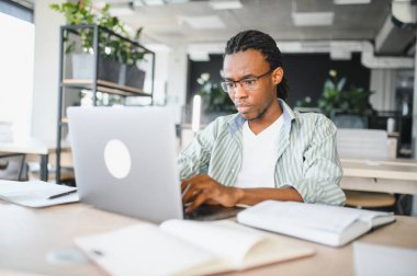 Modern üniversite kütüphanesinde, kitaplar ve notlarla çevrili, dizüstü bilgisayarla çalışan genç bir adam.