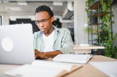 Dizüstü bilgisayarı üzerine yoğunlaşan genç adam, açık kitaplarla, parlak, modern bir kütüphane ortamında, yüksek öğrenimin adanmışlığını somutlaştırıyor.