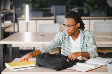 Odaklanmış Afro-Amerikan üniversite öğrencisi kütüphanede çalışırken sırt çantasından bir kitap alıyor.