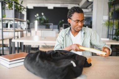 Odaklanmış Afro-Amerikan erkek üniversite öğrencisi modern kütüphanede okurken sırt çantasından ders kitabı alıyor