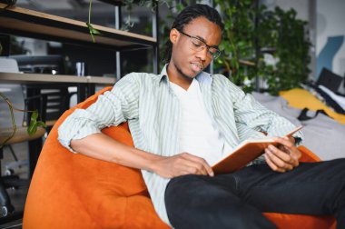 Genç siyahi bir adam, bitki ve kitaplıklarla çevrili modern bir üniversite kütüphanesinde rahat turuncu bir puf üzerinde otururken kitap okuyor.