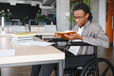 Engelli bir genç tekerlekli sandalye kullanıyor, masasında parlak, modern ofis ortamında kitap okuyor.