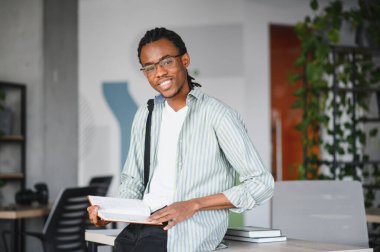 Bilgi ve ilhamla dolu modern bir kütüphanede dururken elinde kitap tutan ve gülümseyen mutlu üniversite öğrencisi.