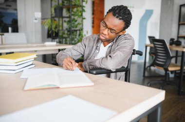 Özürlü genç adam, tekerlekli sandalyeyle deftere yazı yazarak, parlak, modern bir ofiste çalışıyor.