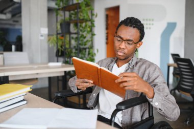 Tekerlekli sandalyedeki genç adam ders kitabını kullanıyor, parlak, erişilebilir ofis alanlarında okuyor, dahil olma ve yüksek öğrenim erişilebilirliğini gösteriyor.