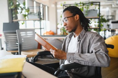 Konsantre olmuş, engelli, tekerlekli sandalye kullanan, parlak, modern bir ofiste çalışırken kitap okuyan ya da çalışan Afro-Amerikan bir adam.