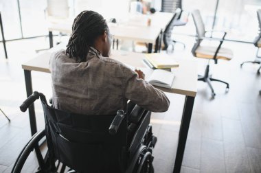 Tekerlekli sandalyede engelli genç bir adam modern bir ofiste not alıyor, çalışıyor ve çalışıyor.