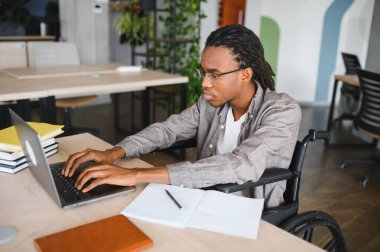 Özürlü genç bir adam, parlak, modern bir ofiste özenle bir dizüstü bilgisayar üzerinde çalışıyor, çalışmalarına odaklanmasını ve kararlılığını somutlaştırıyor.