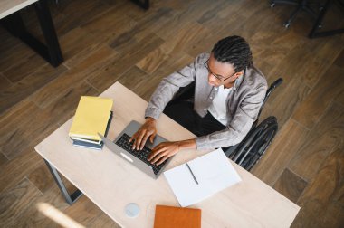Odaklanmış bir Afrikalı Amerikalı öğrencinin, tekerlekli sandalyede otururken dizüstü bilgisayar kullanması, erişilebilirlik, eğitim ve teknoloji temalarını vurgulaması.