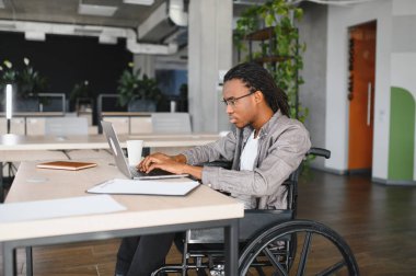 Modern ofiste dizüstü bilgisayarla çalışan engelli genç profesyonel bir adam, iş yerindeki dahil olma ve erişilebilirliği gösteriyor