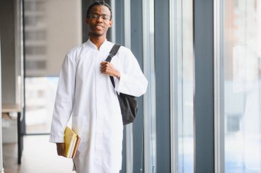 Üniversite koridorunda elinde kitap ve sırt çantasıyla dolaşan kendinden emin bir tıp öğrencisi, derse hazır.