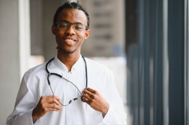 Kendine güvenen genç bir Afro-Amerikan erkek doktorun portresi gülümseyen ve steteskop tutan, sağlık hizmeti profesyonelliği ve uzmanlığını temsil eden