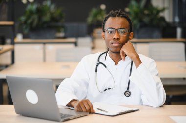 Parlak modern klinikte dizüstü bilgisayar ve tıbbi kayıtlarla uğraşırken düşünceli bir doktor el ele dinleniyor.