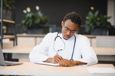 Çağdaş hastane ofisinde masa başında otururken steteskopla not yazan beyaz önlüklü, odaklanmış bir erkek pratisyen.
