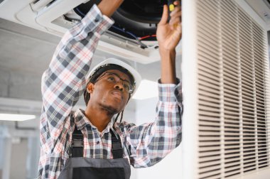Genç Afro-Amerikan erkek teknisyen güvenlik kaskı takıyor ve tulum giyiyor, havalandırma sistemini tornavida kullanarak onarıyor.