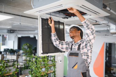 Afrikalı Amerikalı Hvac teknisyeni bir havalandırma ünitesi üzerinde çalışıyor. Modern ofis ortamında bakım ve uygun havalandırmayı sağlıyor.