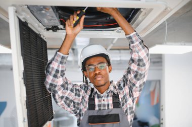 Genç Afro-Amerikan HVK teknisyeni güvenlik başlığı ve gözlük takıyor, havalandırma sistemini tamir ederken tornavida kullanıyor.