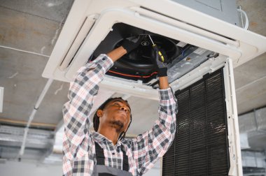 Afrikalı Amerikan Hvac teknisyeni, soğutma sistemi bakımı konusunda uzmanlaşmış bir tornavida kullanarak bir havalandırma ünitesini tamir ediyor.