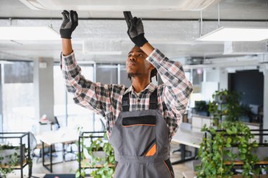 Genç Afro-Amerikan bakım işçisi modern ofiste eldiven ve tulum giyerek havalandırma sistemini tamir ediyor.