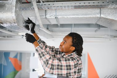 Genç Afrikalı Amerikalı erkek teknisyen ingiliz anahtarı kullanarak modern bir binanın tavanında havalandırma sistemini tamir ediyor.
