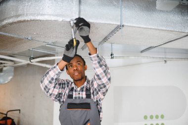 Genç Afrikalı Amerikalı işçi tornavida ve pense kullanarak havalandırma sistemini tamir ediyor, iş tulumu ve eldiven giyiyor.