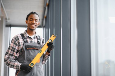 Üniformalı gülen teknisyen elinde havalandırma ve havalandırma tamir işleri için hazır bir aletle bekliyor. Büyük pencerelerin yanında güvenle duruyor
