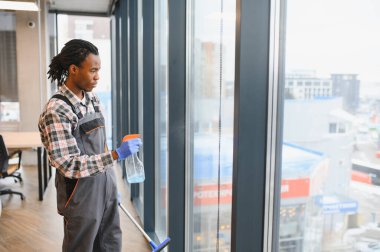 Genç adam, ofis pencerelerini sprey şişesi ve cam sileceği ile temizliyor. Profesyonel görünümü korurken seri bir şekilde parlıyor.