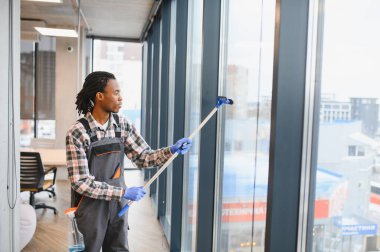 İş tulumu ve eldiven giyen Afrikalı genç bir Amerikalı modern bir ofis binasının camını silmek için cam sileceği kullanıyor.
